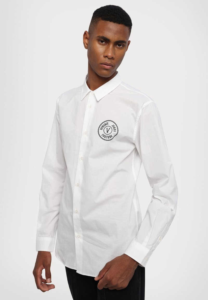 Biała koszula z długim rękawem wykonana z bawełny. Posiada haftowane okrągłe logo na lewej piersi w kolorze czarnym i srebrnym.