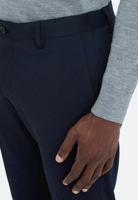 Marineblauwe broek van gestructureerde stof met een slank ontwerp, voorzien van een platte voorkant en knoopsluiting. Hand rustend op het bovenbeen.