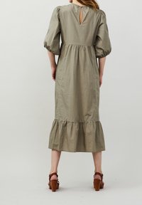 Długa oliwkowo-zielona sukienka z bufkami, z wycięciem z tyłu i falbaniastym dołem. Wykonana z lekkiego materiału z kontrastującymi detalami szwów.