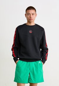 Sort sweatshirt med et geometrisk design, røde detaljer på ærmerne, og et hexagonalt Nike-logo. Sammen med grønne shorts.