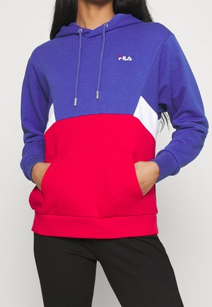 Osoba ubrana w kolorową bluzę z kapturem FILA w kolorach niebieskim, czerwonym i białym, z przednią kieszenią, ręce w kieszeniach oraz widoczne czarne spodnie.