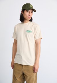 Hombre con gorra oliva de Nueva York, camiseta beige con el logo verde "OBEY" y pantalones de pana marrones, de pie frente a un fondo blanco liso.