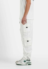 Witte cargo-broeken met vier zakken en zwarte vierkante sluitingen, gecombineerd met grijs-groene sneakers, gezien vanaf de zijkant.