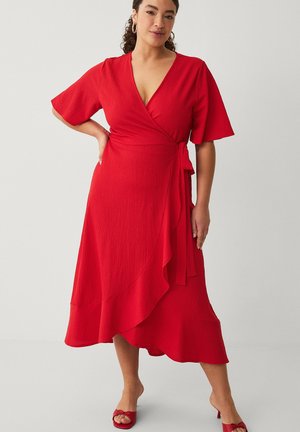 Kvinde står med hånden på hoften iført en rød slå-om-kjole med flagrende ærmer og flæsekant, kombineret med røde åbne tå-sandaler.