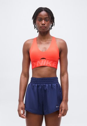 Jonge vrouw staand, met een oranje sportbeha en marineblauwe korte sportbroek, tegen een effen lichte achtergrond.