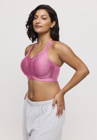 Reggiseno sportivo rosa con motivo strutturato, spalline larghe e design di supporto. Indossato con pantaloni della tuta grigi, evidenziando una vestibilità aderente e comfort.