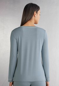 Ljusblå långärmad topp med rund hals, som har en mjuk textur och kontrasterande vit sömnad längs halsen och ärmsluten.