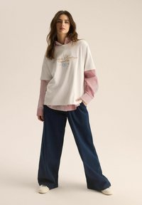 Biała, oversize'owa koszulka z złotym haftem nałożona na czerwoną i białą w pasy koszulę, w zestawieniu z ciemnymi spodniami o szerokich nogawkach i białymi butami.