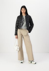 Doudoune noire, design court, ajustée à la taille. T-shirt graphique gris, pantalon beige amples, sac blanc et chaussures de sport.
