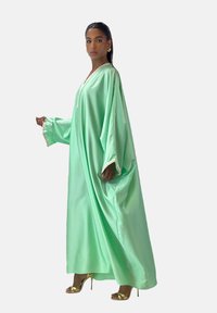 Abito lungo in raso verde con maniche larghe, scollo a V e rifiniture decorative. La modella indossa tacchi dorati, mettendo in risalto una silhouette fluida e oversize.