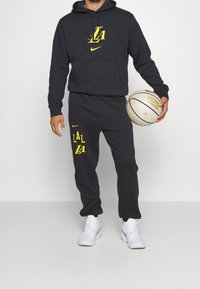 Sort hættetrøje og sweatpants sæt med gul "LA" grafik. Indeholder en kængurulomme, Nike-logo og er parret med hvide atletiske sko og en basketball.