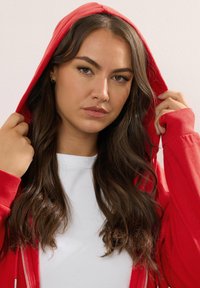 Sudadera roja con cremallera y capucha forrada, llevada sobre una camiseta blanca. La tela parece suave y el diseño presenta puños acanalados.