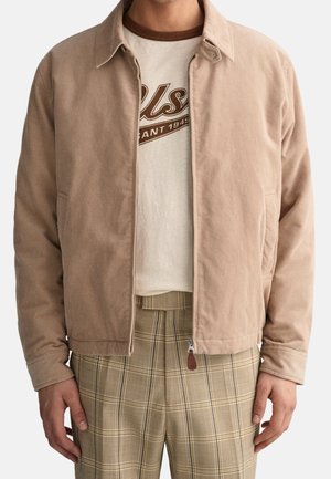 Mężczyzna w beżowej sztruksowej kurtce na zamek, kremowym t-shircie z grafiką oraz beżowych spodniach w kratę, widoczny od ramion do bioder.