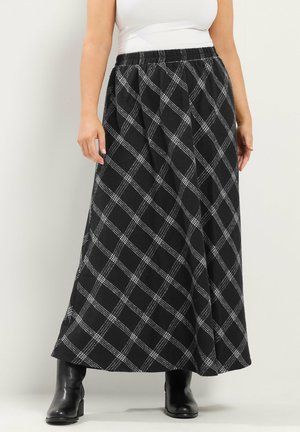 Vrouw die een lange zwart-witte geruite rok draagt, zwarte enkellaarsjes en een witte mouwloze top, staand tegen een effen muur.