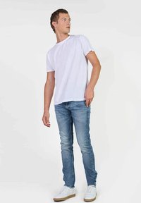 T-shirt bianco in cotone abbinato a jeans azzurri chiari. Modella che indossa sneakers bianche, in posizione neutra su uno sfondo semplice.