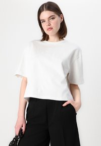 Top alb cu mâneci scurte, cu un croi lejer, având modele de cusături vizibile și un guler rotund, combinat cu pantaloni negri.