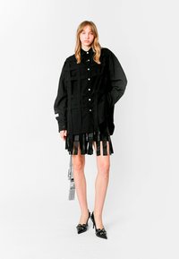 Camisa oversized negra con detalles de flecos, cierre delantero con botones y acentos bordados. La modelo lleva una falda corta y tacones altos negros.