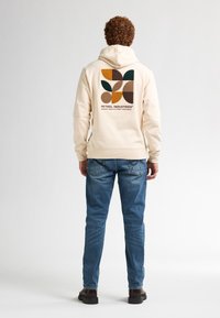 Sudadera color crema con un diseño de hojas abstractas y coloridas en la parte posterior; combinada con jeans azules y botas marrones. Textura de tela suave.