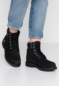 Schwarze Wildleder-Ankle Boots mit runder Zehenpartie, schwarzen Schnürsenkeln, türkisfarbenen Ösen und einem glänzenden schwarzen Absatz; strukturierte Gummisohle, kombiniert mit blauen Jeans.