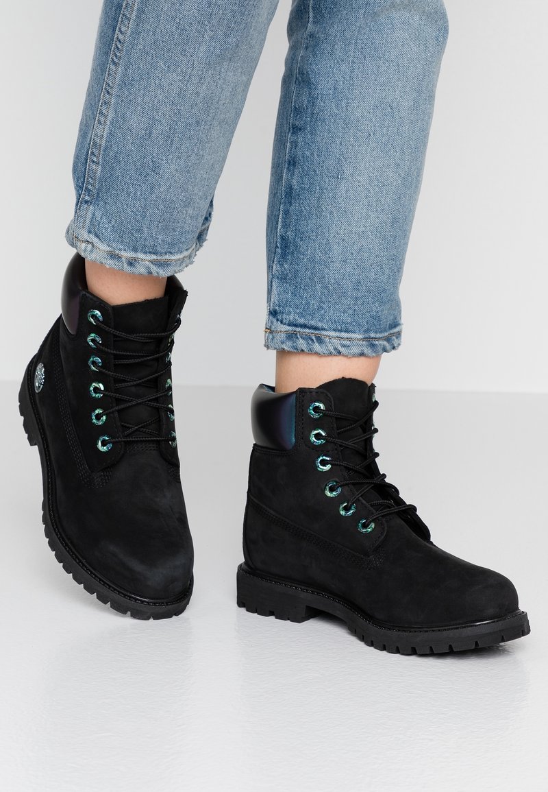 Schwarze Wildleder-Ankle Boots mit runder Zehenpartie, schwarzen Schnürsenkeln, türkisfarbenen Ösen und einem glänzenden schwarzen Absatz; strukturierte Gummisohle, kombiniert mit blauen Jeans.