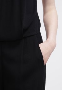 Combinaison noire en tissu texturé ; présente un haut ample et un pantalon ajusté avec des poches latérales, mettant en valeur un design minimaliste.