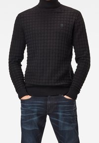 Svart polotröja med ett strukturerat rutmönster, ribbade muddar och fåll. Bärs med mörkblå jeans, händerna i fickorna.