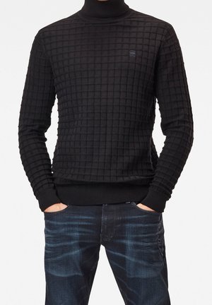 Schwarzer Rollkragenpullover mit einem strukturierten Rautenmuster, gerippten Bündchen und Saum. Tragen mit dunkelblauen Jeans, Hände in den Taschen.