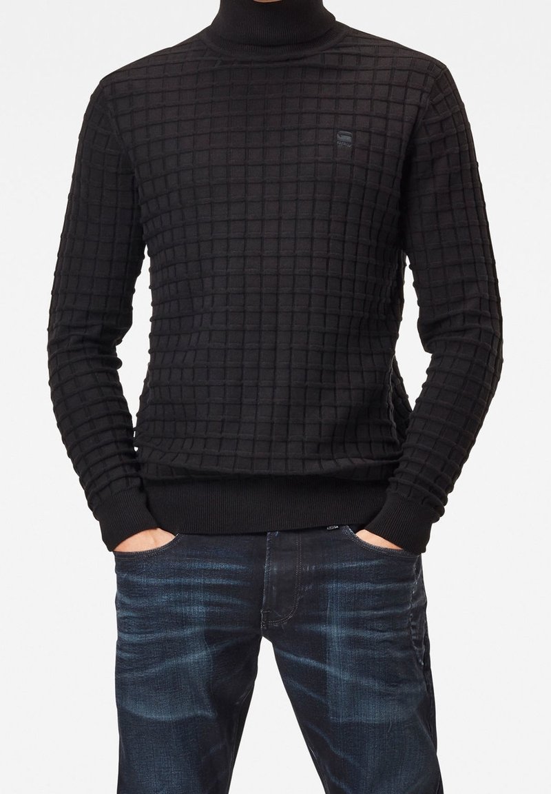 Svart polotröja med ett strukturerat rutmönster, ribbade muddar och fåll. Bärs med mörkblå jeans, händerna i fickorna.