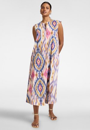 Kvinde stående iført ærmeløs, ankel-længde kjole med farverigt geometrisk mønster og gyldne remsandaler på ensfarvet baggrund.