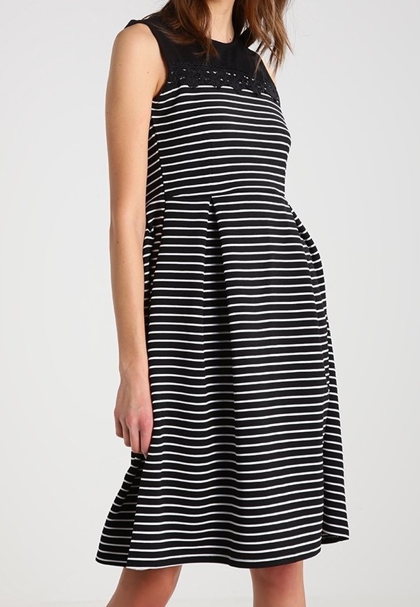 Ärmlös svart klänning med vita horisontella ränder, spetsdetalj på bröstet och plisserad kjol, bärare står upp.