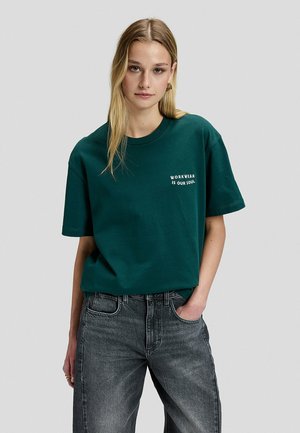 Junge Frau mit langen blonden Haaren trägt ein grünes T-Shirt mit der Aufschrift "Workwear is our soul" und eine graue Jeans, stehend vor einem schlichten hellen Hintergrund.
