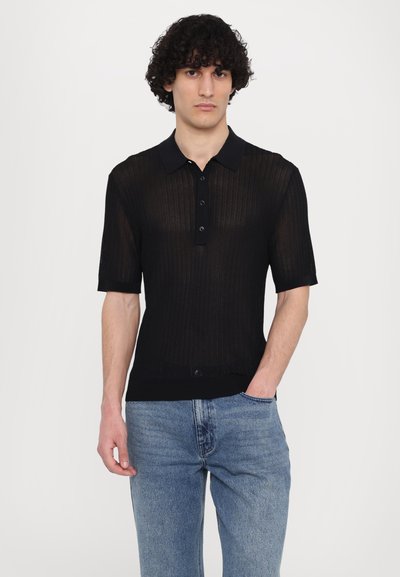 Fekete rövid ujjú polo póló, textúrált bordázott mintával, galléros nyakkal és négy gombos kapoccsal. Kék farmernadrággal viselve.