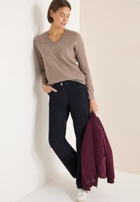 Vaaleanruskea v-kaula-aukollinen villapaita, tummansiniset farkut hienovaraisilla raidallisilla kuvioilla, viininpunainen bomber-takki kädessä, valkoiset lenkkarit, neutraali tausta.