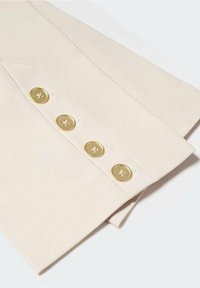 Manchette en tissu beige avec quatre boutons dorés uniformément espacés cousus en ligne droite près du bord du poignet.