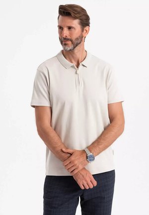 Lys beige polo-shirt med korte ærmer og en lynlåskrage, lavet af blødt stof; parret med mørkt ternede bukser.