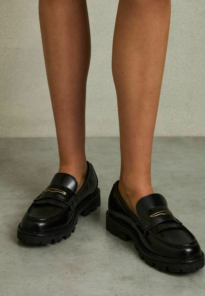 Sorte læderloafer med en tekstureret finish, ribbet gummisål og guldaccent over vampen. Har et traditionelt frynse-design.