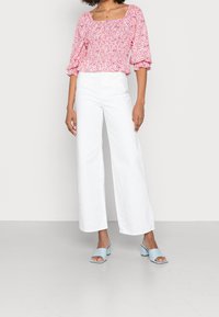 Top floreale con maniche a sbuffo rosa su sfondo bianco, abbinato a pantaloni bianchi a vita alta e gamba larga e sandali con tacco a blocco blu chiaro.