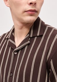 Άνδρας που φοράει καφέ πουκάμισο με κάθετες λευκές ρίγες, αποκομμένο από τον λαιμό μέχρι το μέσο του στήθους, κοιτάζοντας προς το πλάι.