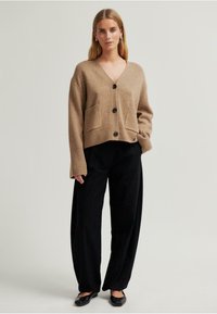 Beige kötött kardigán V nyakkivágással, hosszú ujjal és elülső zsebekkel, fekete gombokkal és bordázott mandzsettával, fekete nadrággal párosítva.