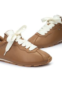 Baskets en cuir tan avec une pointe arrondie, des lacets blancs, des coutures décoratives et de petits accents métalliques sur le côté.