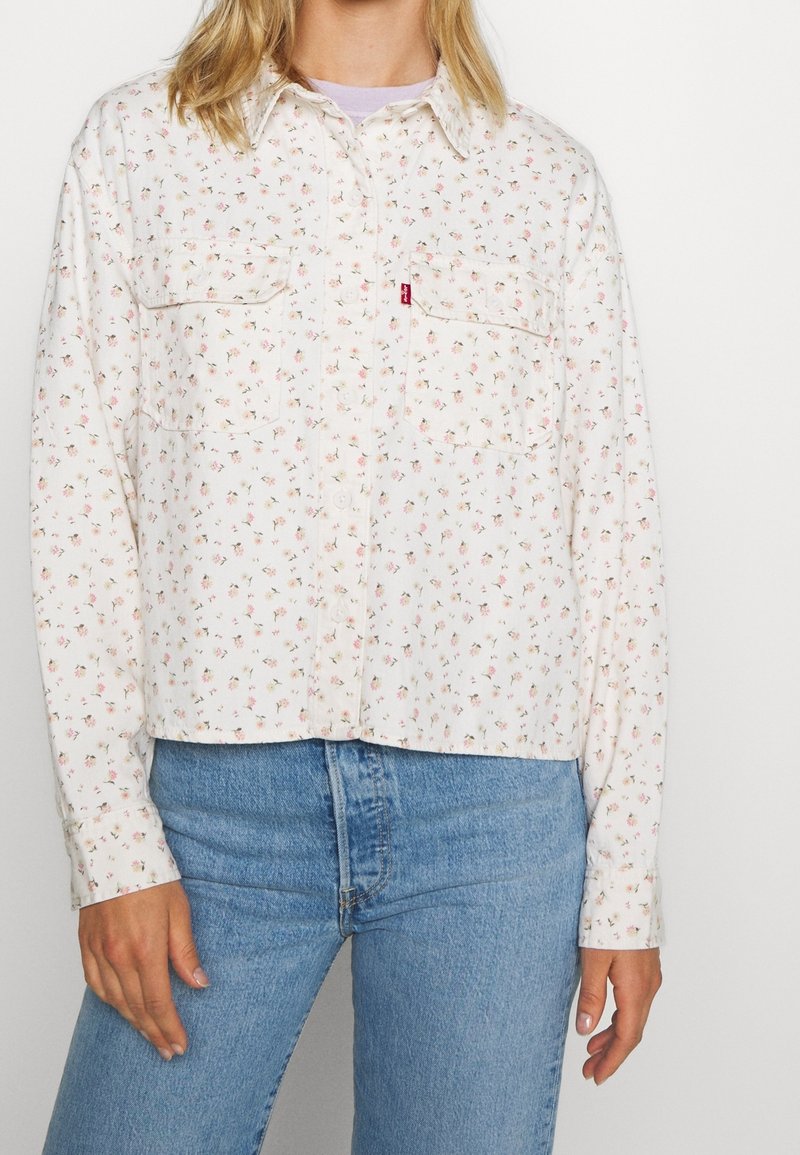 Chemise blanche à manches longues avec petit motif floral rose, boutonnée sur le devant, deux poches poitrine et col pointu. Associée à un jean bleu.