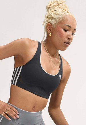 Jeune femme aux cheveux blonds bouclés, portant un brassière de sport noire Adidas et un legging gris, penchée en avant avec une expression concentrée.