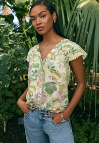 Bluse med blomstermønster og korte ærmer i lys gul med grønne detaljer, med v-halsudskæring og knapper, parret med lyseblå denimjeans.