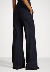 Pantaloni a gamba larga a righe navy con vita alta, dotati di tasche laterali e una texture liscia. Il taglio sartoriale valorizza la silhouette.
