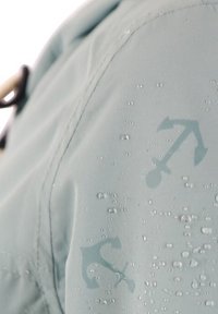 Vattentålig ljusblå jacka av tyg med synliga vattendroppar och subtilt ankarmönster på axeln.