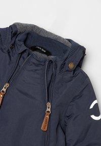 Veste bleu marine avec fermeture éclair à l'avant et capuche, avec tirettes de fermeture éclair en cuir, bouton-pression au col et doublure polaire grise à l'intérieur de la capuche.