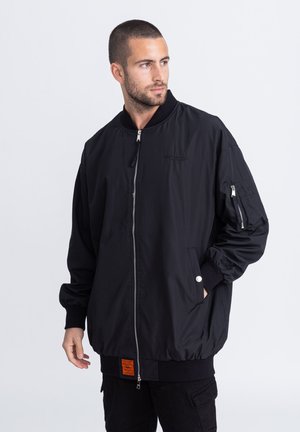 Czarna kurtka bombowiec z gładkiego materiału, z zamkiem błyskawicznym z przodu, bocznymi kieszeniami, ściągaczami na mankietach oraz pomarańczową metką na dole.