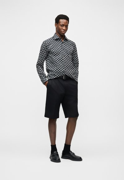 Jeune homme debout, les mains dans les poches, portant une chemise à manches longues à motifs noir et blanc, un short noir, des chaussettes noires et des mocassins noirs.
