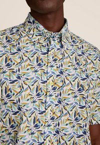 Korte mouwen overhemd met knopen in witte stof met een meerkleurig bladenpatroon in groen, blauw en geel. Heeft een spreidkraag en gele knopen.