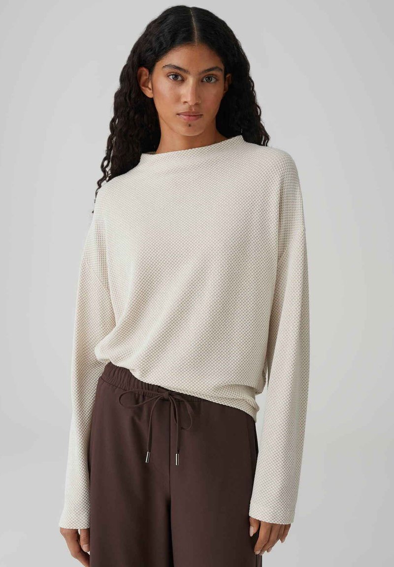 Haut à manches longues beige clair avec tissu texturé, coupe décontractée et encolure large, associé à un pantalon marron foncé à cordon de serrage.